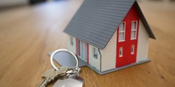 A toy house on a table, with gre roof and red and white walls. A door key is laying next to it.
