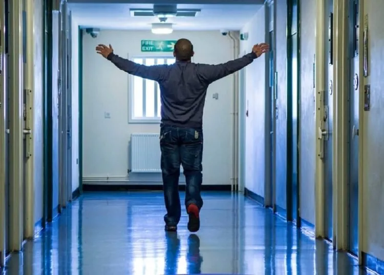 A man standing in a corridor with doors to both sides. He has his back to us, and his arms stretched out to the sides.