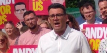 Man in a white polo shirt speaks at a political rally as a crowd behind him holds bright pink signs reading 'ANDY FOR US'.