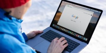 Person in blue jacket and red cap uses laptop and has google search open